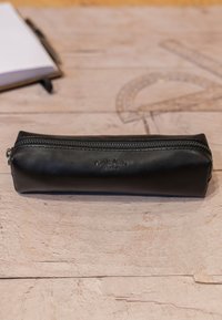 Zwarte leren etui met een soepele textuur, voorzien van een ritssluiting aan de bovenkant en het reliëfmerk aan de zijkant. Rechthoekige vorm.