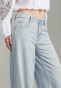 Jeans estilo wide-leg de denim azul claro con una cintura alta, que presentan bolsillos en la parte posterior y una sutil textura desgastada.