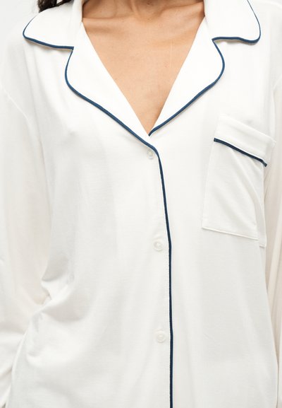 Haut de pyjama blanc à boutons avec passepoil bleu marine le long du col, de la poche et de la patte de boutonnage avant, porté par une personne.