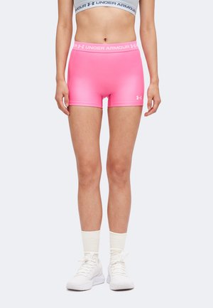 Nő élénk rózsaszín Under Armour kompressziós rövidnadrágban, fehér sportmelltartóban, fehér zokniban és fehér sportcipőben, egyszínű háttér előtt áll.