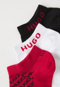 Trys poros kulkšninių kojinių sukrautos, vienos juodos ir baltos su "HUGO" tekstu, vienos raudonos su juodu pakartotiniu "HUGO" raštu.