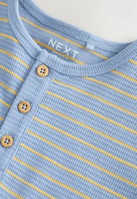 Hele sinine tekstuuriga kangast T-särk kollaste triipudega, ümmarguse kaelusega ja kolme puidust nupuga krae ees. Bränd "NEXT" on nähtav.