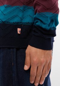 Pull en tricot avec un motif texturé dans des teintes bordeaux, sarcelle et marine. Présente une étiquette décorative à l'ourlet et des bords côtelés.