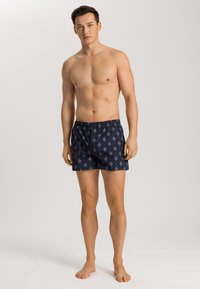Boxer in blu navy con un motivo a rombi ripetitivo in rosso chiaro e bianco. Vita elasticizzata, vestibilità ampia, indossati a piedi nudi.