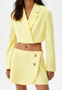 Yellow cropped blazer with a lapel collar and two-button closure, paired with a matching wrap skirt featuring a side button detail.