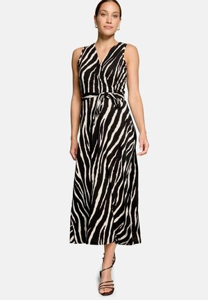 Femme portant une robe portefeuille sans manches à imprimé zèbre noir et blanc avec une taille nouée, associée à des sandales noires à brides et des boucles d'oreilles créoles dorées.