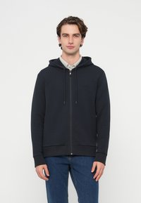 Námornícka modrá mikina na zips s kapucňou so šnúrkou, rebrovanými manžetami a lemom. Mäkká bavlnená zmes. Model má pod ňou svetlú košeľu s golierom.