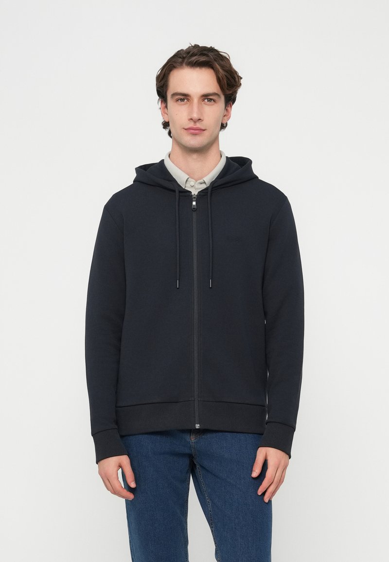 Námornícka modrá mikina na zips s kapucňou so šnúrkou, rebrovanými manžetami a lemom. Mäkká bavlnená zmes. Model má pod ňou svetlú košeľu s golierom.