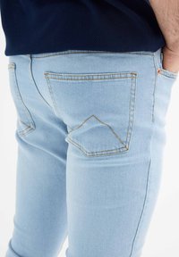 Jeans de mezclilla azul claro con un ajuste slim, bolsillos traseros estándar con solapas puntiagudas y detalle de costura amarilla en contraste.