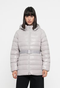 Sivá vyteplená bunda s kapucňou a predným zipsom. Obsahuje opásaný pás a lesklú textúru, s nápisom "Calvin Klein" na hrudi a opasku.