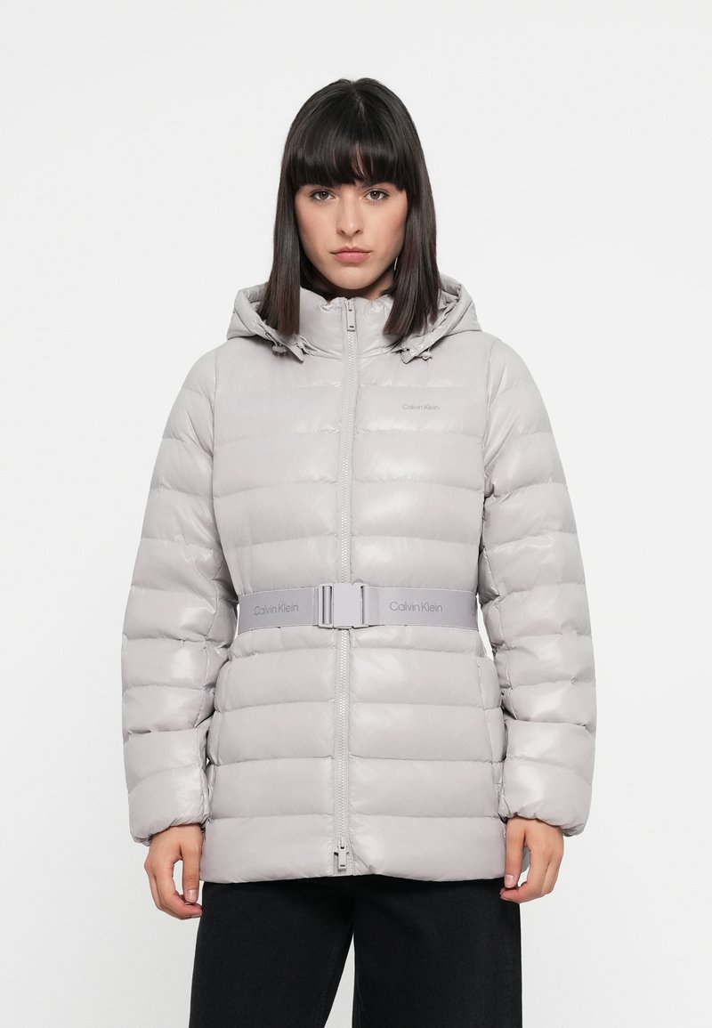 Grijze gewatteerde jas met een capuchon en een ritssluiting aan de voorkant. Voorzien van een ceintuur in de taille en een glanzende textuur, gekenmerkt met "Calvin Klein" op de borst en de ceintuur.