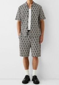 Hombre vestido con una camisa de manga corta de patrón geométrico en blanco y negro y pantalones cortos a juego, con calcetines blancos y zapatos negros, de pie contra una pared blanca.