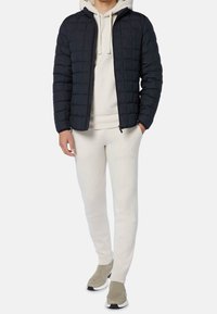 Svart kviltad puffjacka med dragkedja framtill, bärs över en ljus hoodie och krämfärgade joggers, tillsammans med beige sneakers.