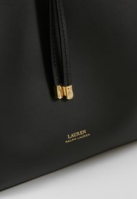 Sac à main en cuir noir avec deux Zips dorés, arborant une marque métallisée "LAUREN RALPH LAUREN". Texture lisse, design minimal, sans motifs visibles.