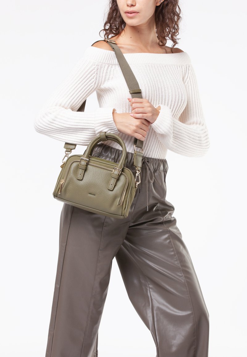 Vrouw in een off-shoulder witte gebreide trui en bruine leren broek, met een olijfgroene leren handtas met handvatten aan de bovenkant en een schouderband.
