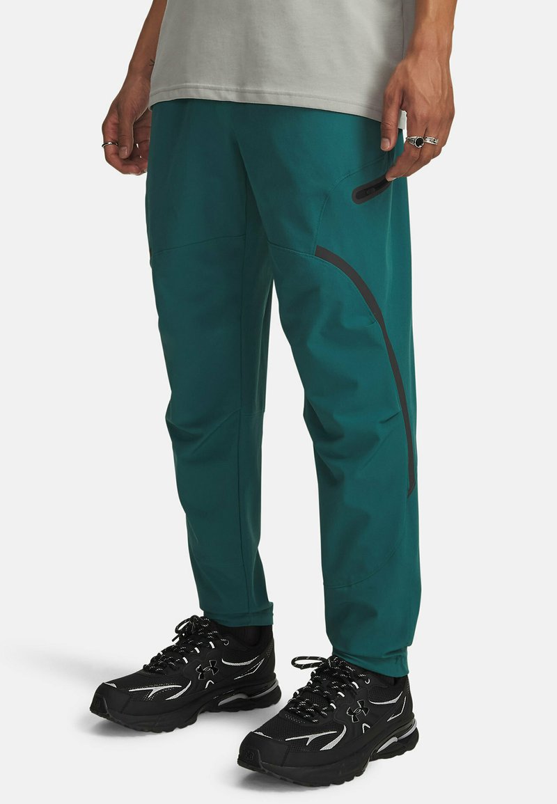 Teal friluftsbukser lavet af letvægtsmateriale, med sorte accenter, lynlåslommer og en tilspidset pasform. Kombineret med sorte atletiske sko.