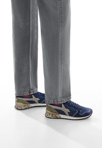 Sneakers atletici blu e grigi con una suola a motivo camouflage, dotati di un accento beige e un interno rosso. Indossati con jeans grigi.