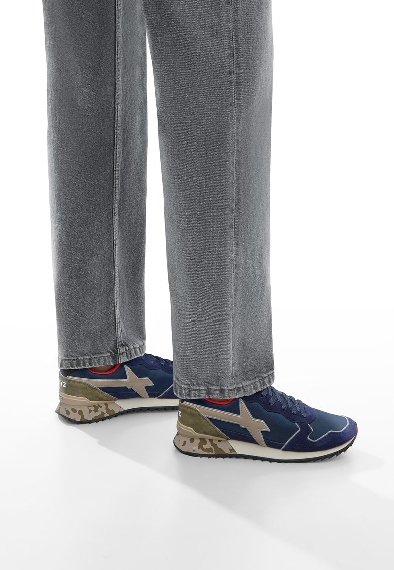 Sneakers atletici blu e grigi con una suola a motivo camouflage, dotati di un accento beige e un interno rosso. Indossati con jeans grigi.