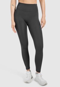 Leggings noires taille haute avec une texture lisse et extensible, un design ajusté et des coutures renforcées. Portés avec des chaussures de sport blanches.