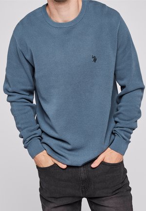 Homme portant un pull bleu texturé à manches longues avec un petit logo noir sur le côté gauche de la poitrine, mains dans les poches, associé à un jean noir.