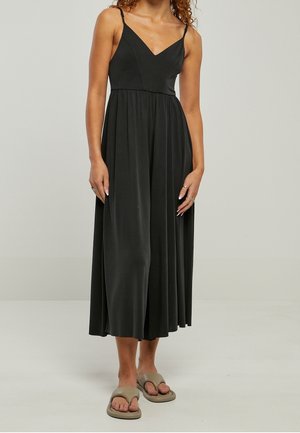 Vrouw draagt een zwarte mouwloze midi-jurk met een V-hals en geplooide rok, gecombineerd met beige slip-in sandalen.