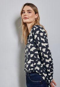 Blusa a maniche lunghe di colore blu navy, con motivi astratti bianco sporco. Il tessuto sembra leggero con una vestibilità ampia e un collo rotondo.