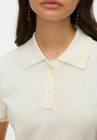 Polówka w kolorze kremowym o miękkiej fakturze, z kołnierzykiem i plisą z sześcioma jasnymi guzikami. Zbliżenie na linię dekoltu.