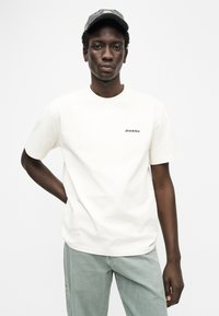 Εκρού βαμβακερό t-shirt με κοντά μανίκια, στρογγυλή λαιμόκοψη και μαύρο κεντητό λογότυπο «Dickies» στο αριστερό στήθος. Συνδυάζεται με ανοιχτοπράσινο παντελόνι.