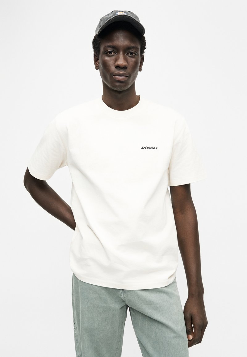 Crèmekleurig katoenen T-shirt met korte mouwen, ronde hals en zwart geborduurd "Dickies"-logo op de linkerborst; gecombineerd met lichtgroene broek.