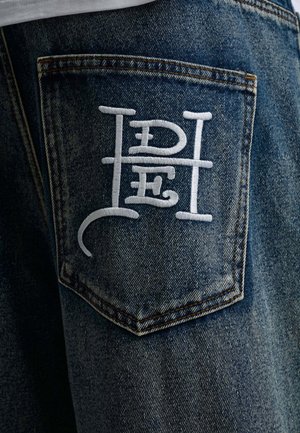 Lynlomme i denimjeans med hvide broderede stiliserede bogstaver, der danner et unikt monogramdesign på bagsiden.