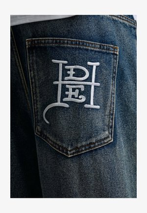 Poche de jean en denim avec des lettres stylisées blanches brodées formant un monogramme unique au dos.