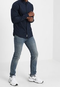 Granatowa koszula na guziki, jasnoniebieskie dżinsy i białe trampki. Prosty design o gładkiej fakturze i dopasowanym kształcie.