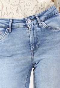 Ljust blå denimjeans med dragkedja och knappstängning, fem fickor och synliga s ömmar. Slät textur.