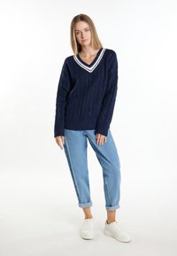Marineblaue Strickpullover mit V-Ausschnitt, der weiße Streifen entlang des Ausschnitts aufweist. Kombiniert mit hellblauen, schmalen Jeans und weißen Sneakers.
