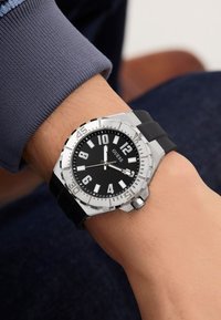 Orologio con quadrante nero, numeri bianchi ben visibili, cassa in acciaio inossidabile argentato e cinturino in gomma nera texturizzata. Presenta una lunetta lucidato.