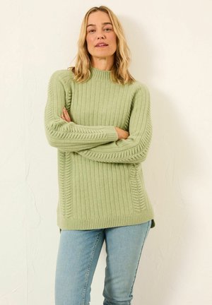 Hellgrüner Strickpullover mit geripptem Muster, hohem Kragen und seitlichen Details. Kombiniert mit hellblauen Jeans.