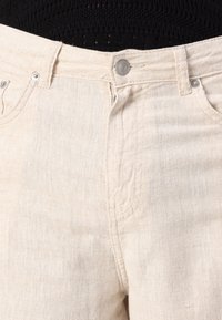 Jeans bej deschis cu o textură subtilă. Prezintă un sistem de închidere cu un buton metalic, bucle pentru brâu și un design standard cu cinci buzunare.