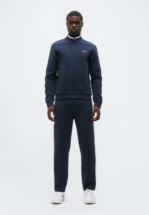 Muž stojaci, oblečený v tmavomodrej teplákovej súprave so zipsovou bundou a zodpovedajúcimi nohavicami, doplnený bielymi teniskami, na jednoduchom svetlom pozadí.