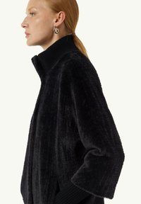 Maglione in peluche nero con un alto colletto, polsini a coste e una vestibilità morbida. Il tessuto ha una texture morbida con righe verticali.