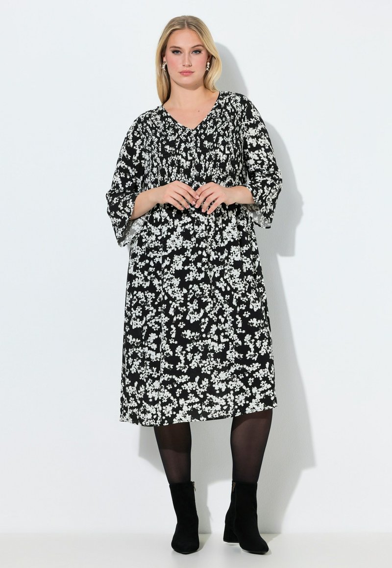 Vestito nero a fiori con motivi bianchi, scollo a V, maniche larghe, lunghezza a metà polpaccio, indossato con stivaletti neri. Tessuto dalla texture liscia.