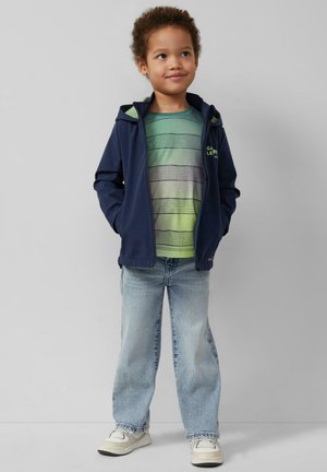 Jeune garçon debout, les mains dans les poches, portant une veste à capuche bleu marine, une chemise rayée verte, un jean bleu clair et des baskets blanches, sur un fond uni.