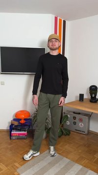 Black long-sleeve shirt, olive green pants, and gray sneakers. Standing in a room with a television, colorful wall, and decorative elements.
