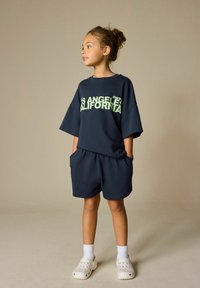 Tmavomodré tričko z bavlny s nadrozmernými rukávmi a svetlozeleným nápisom "Los Angeles California"; zladené šortky s vreckami a biele slip-on topánky.