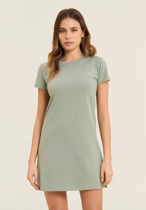 Mujer joven con cabello castaño claro vistiendo un vestido casual de manga corta color verde salvia, de pie frente a un fondo beige liso.