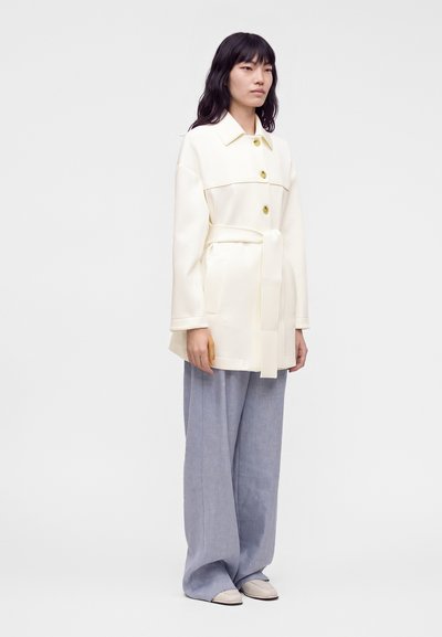 Femme aux longs cheveux noirs portant un manteau ceinturé écru avec de gros boutons et un pantalon ample gris clair sur un fond uni.