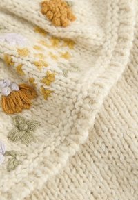 Tissu en tricot beige avec des broderies texturées dans des motifs floraux orange, jaune, vert et violet ; aspect doux et chaud.