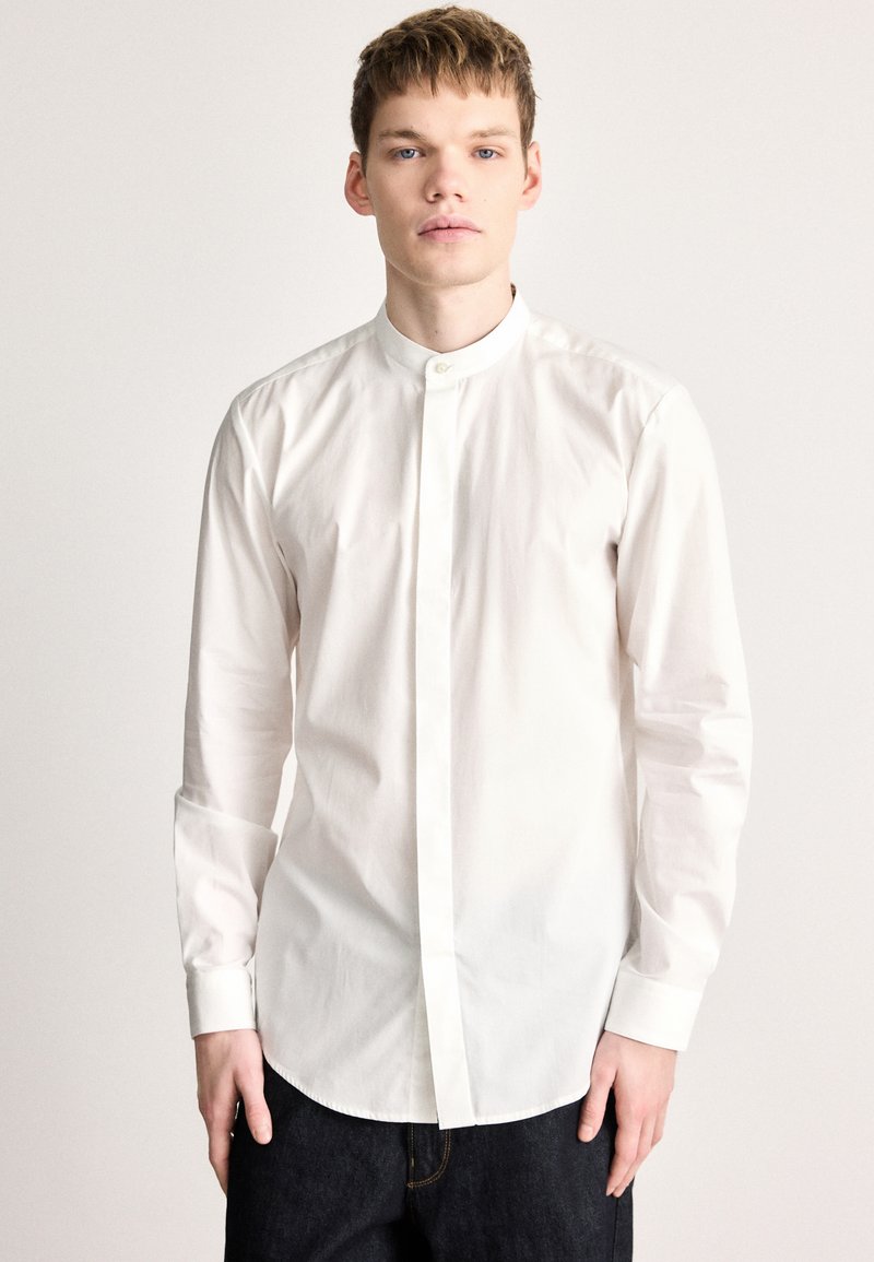 Άσπρο πουκάμισο μακρυμάνικο με λαιμόκοψη τύπου μπαντάνα, κατασκευασμένο από απαλό ύφασμα. Διαθέτει ίσια βάση και κλείσιμο με κουμπιά στο λαιμό.