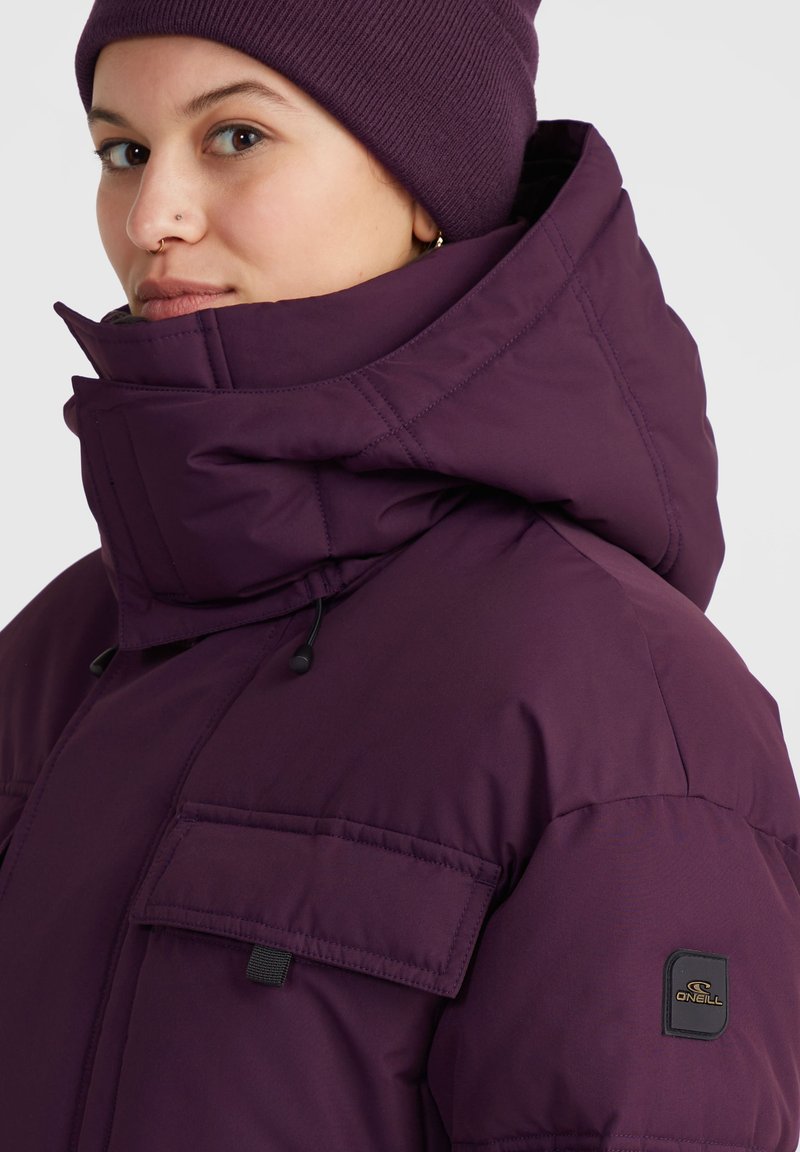 Lila isolierte Jacke mit hohem Kragen, großen Fronttaschen, Kordelzügen und einem Logopatch am Ärmel. Dazu eine passende Mütze.