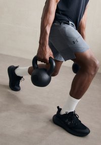Juodas kettlebell, pažymėtas 12KG, laikomas viena ranka, dėvimi pilki sportiniai šortai, juodi sportbačiai ir balti kojinai. Dėmesys raumeningai rankai ir suėmimui.