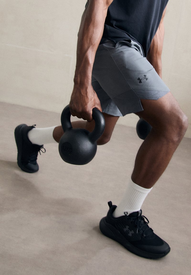 Juodas kettlebell, pažymėtas 12KG, laikomas viena ranka, dėvimi pilki sportiniai šortai, juodi sportbačiai ir balti kojinai. Dėmesys raumeningai rankai ir suėmimui.